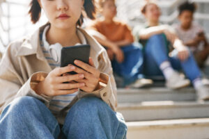 Closeup of teenage girl holding smartphone outdoors while sitting on metal stairs with group of friends in background, copy space - Social Media Lawsuit - Social Media Lawyer - Harding Mazzotti, LLP